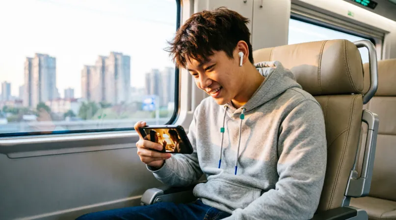 A young man on a train smiling at a film playing on his phone, with earbuds in and the city passing outside